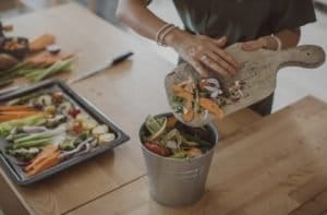 A woman is brushing kitchen scraps off of a cutting board into a bucket